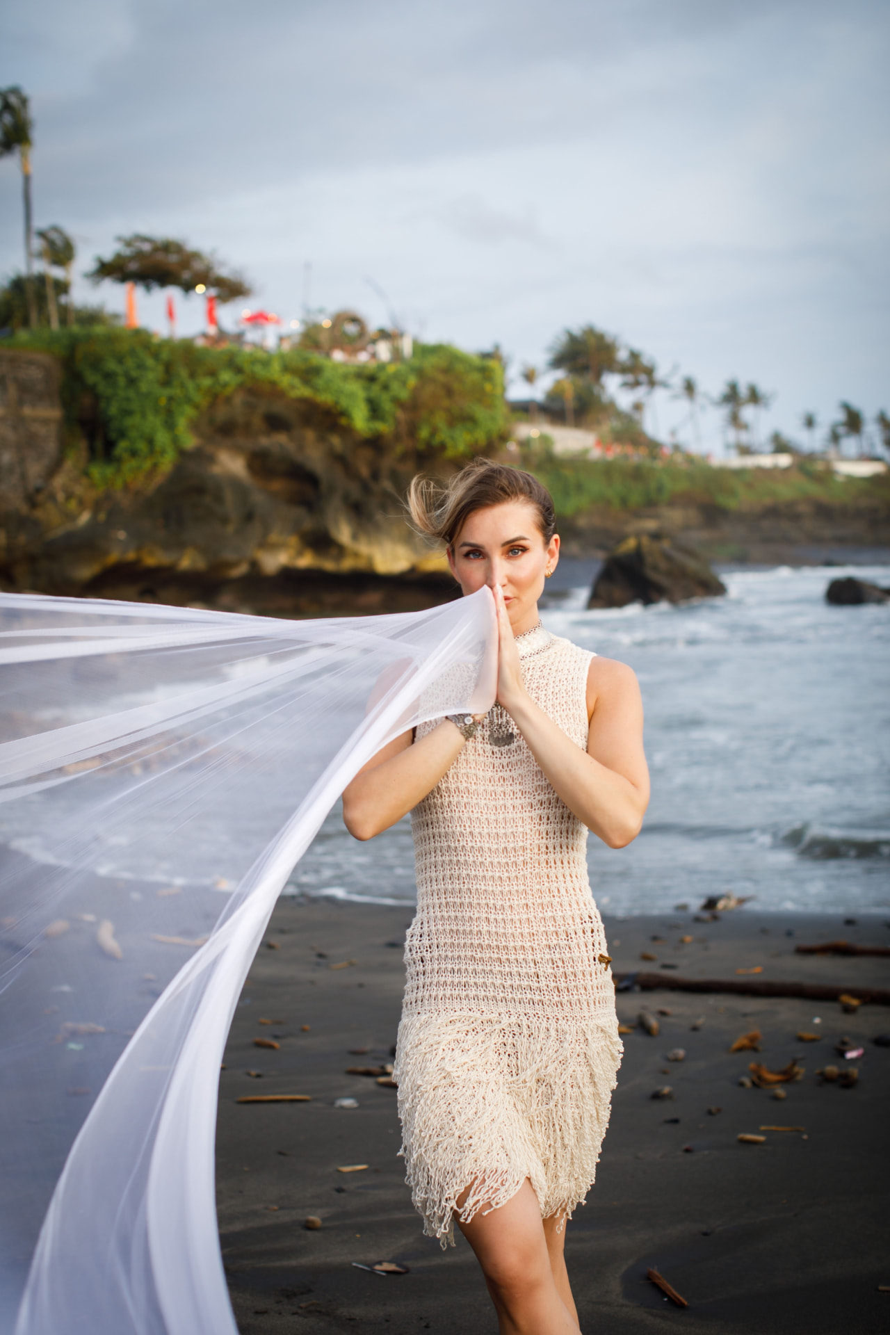 Monika standing on a black sand beach, hands pressed together, white fabric flowing beside her